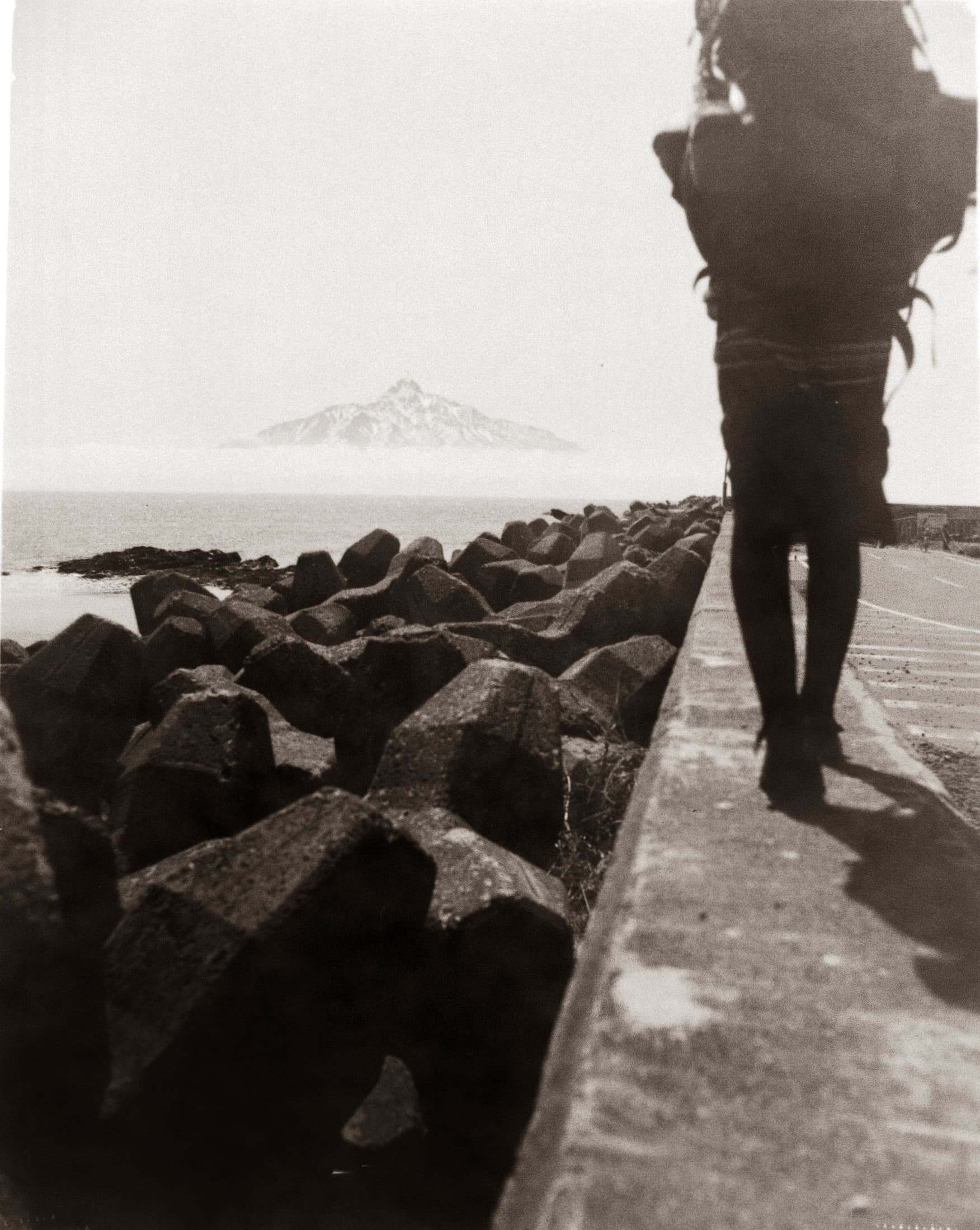 Black-and-white photo of a hiker with a large pack walking along a concrete path beside coastal tetrapods, with a mountain visible through mist in the distance.